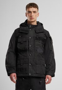 Giacca tattica nera con molteplici tasche, cappuccio regolabile e toppe in Velcro. Realizzata in tessuto resistente con una finitura strutturata.