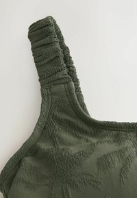 Gros plan sur un tissu vert foncé texturé avec des motifs de feuilles de palmier en relief sur un vêtement à bretelle froncée.