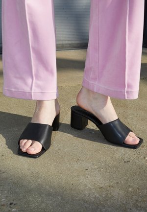 Persona con pantalones rosas y sandalias negras de tacón ancho y apertura en los dedos, de pie sobre una superficie de hormigón, con un talón levantado.
