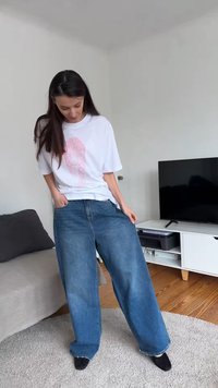 Sieviete valkā pārlielus zilos džinsas un baltu T-kreklu ar rozā dizainu, stāv viesistabā pie TV un dīvāna, skatās uz leju un tur džinsas.