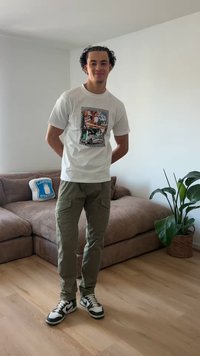 Jonge man staat binnen bij een bruine bank met een Lawrence Station kussen en een grote groene kamerplant op een houten vloer.