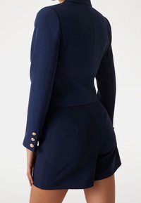 Marineblauwe op maat gemaakte blazer met een aansluitend ontwerp, lange mouwen en gouden knoopaccenten. Gepaarde met bijpassende shorts, die een gestroomlijnde silhoutte tonen.