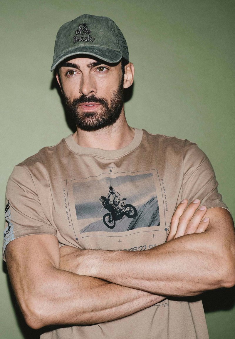 Beżowy t-shirt z grafiką przedstawiającą motocykl, krótki rękaw i okrągły dekolt. Oliwkowa czapka z haftowanym logo. Model pozuje z założonymi rękami.