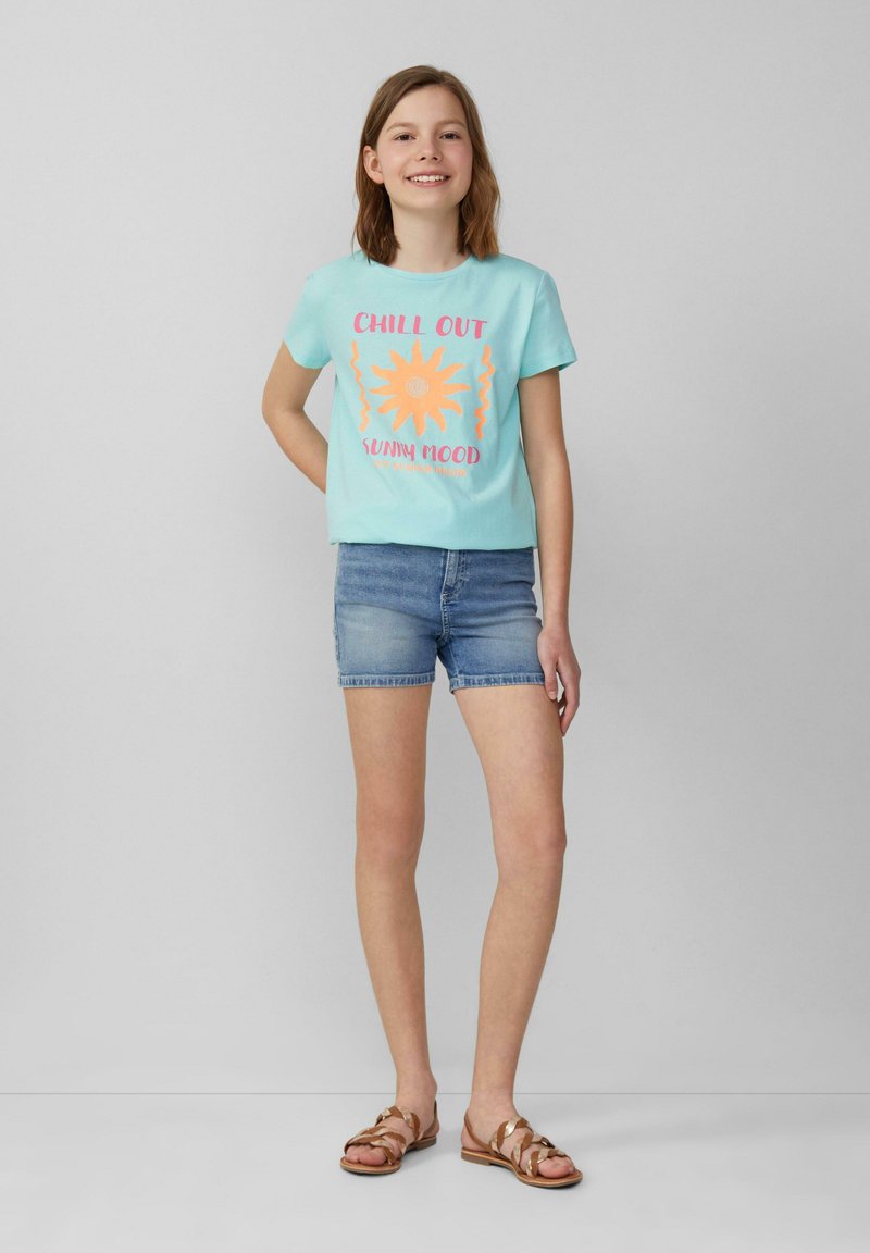 Hellblaues T-Shirt mit dem Text "CHILL OUT SUNNY MOOD" und einer Sonnenillustration. Kombiniert mit Jeansshorts und braunen Sandalen mit Riemen auf einem schlichten Hintergrund.