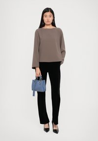Haut marron à manches longues, pantalon en velours noir et escarpins noirs à bout pointu. Tenant un petit sac à main bleu. Fond blanc uni.