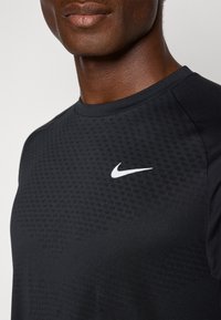 Sort atletisk skjorte lavet af åndbart stof, med et struktureret mønster, rund halsudskæring og et hvidt Nike-logo på brystet.