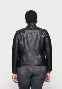 Černá kožená bunda s přiléhavým střihem, se stojatým límcem a švy. Stylizováno s tmavě šedými džíny, zobrazeno zezadu.