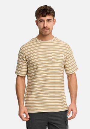 Mężczyzna ubrany w beżowo-biały, poziomo paskowany t-shirt z krótkim rękawem i kieszonką na piersi, stojący na jednolitym tle.