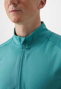 Mėlyna zip-up striukė su aukštu apykakle, pasižyminti sklandžiu paviršiumi, matomomis siūlėmis ir subtilia užtrauktuko detale prie kaklo.