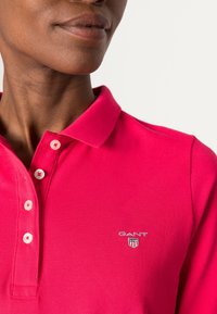 Růžová polo tričko s texturovaným povrchem, se dvěma knoflíky na léze a malým vyšitým logem na levé hrudi.