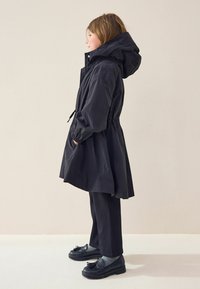 Parka neagră impermeabilă cu glugă ajustabilă, talie strânsă, buzunare laterale, asortată cu pantaloni negri și încălțăminte cu un design texturat.