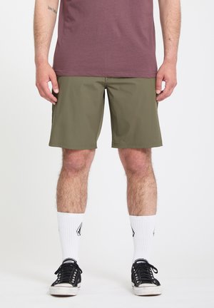 Homme portant un short vert olive, des chaussettes blanches avec des logos noirs, des baskets noires et un t-shirt marron, debout sur un fond blanc.