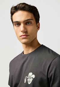 Camiseta negra de cuello redondo y mangas cortas, con un gráfico de un vaquero de dibujos animados en blanco en el lado izquierdo del pecho. Textura suave.
