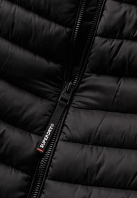 Giubbotto imbottito nero con texture trapuntata, dotato di zip frontale e un'etichetta in tessuto "SUPERDRY" con un accento rosso.