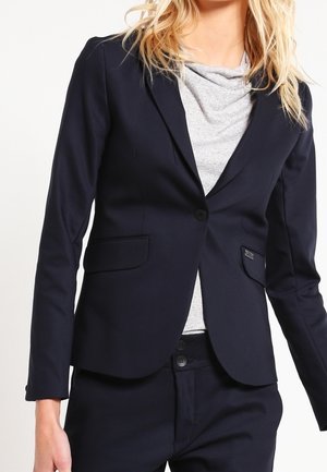 Mujer que lleva una blazer azul marino ajustada con un botón, sobre una camiseta gris claro, combinada con pantalones azul marino a juego.