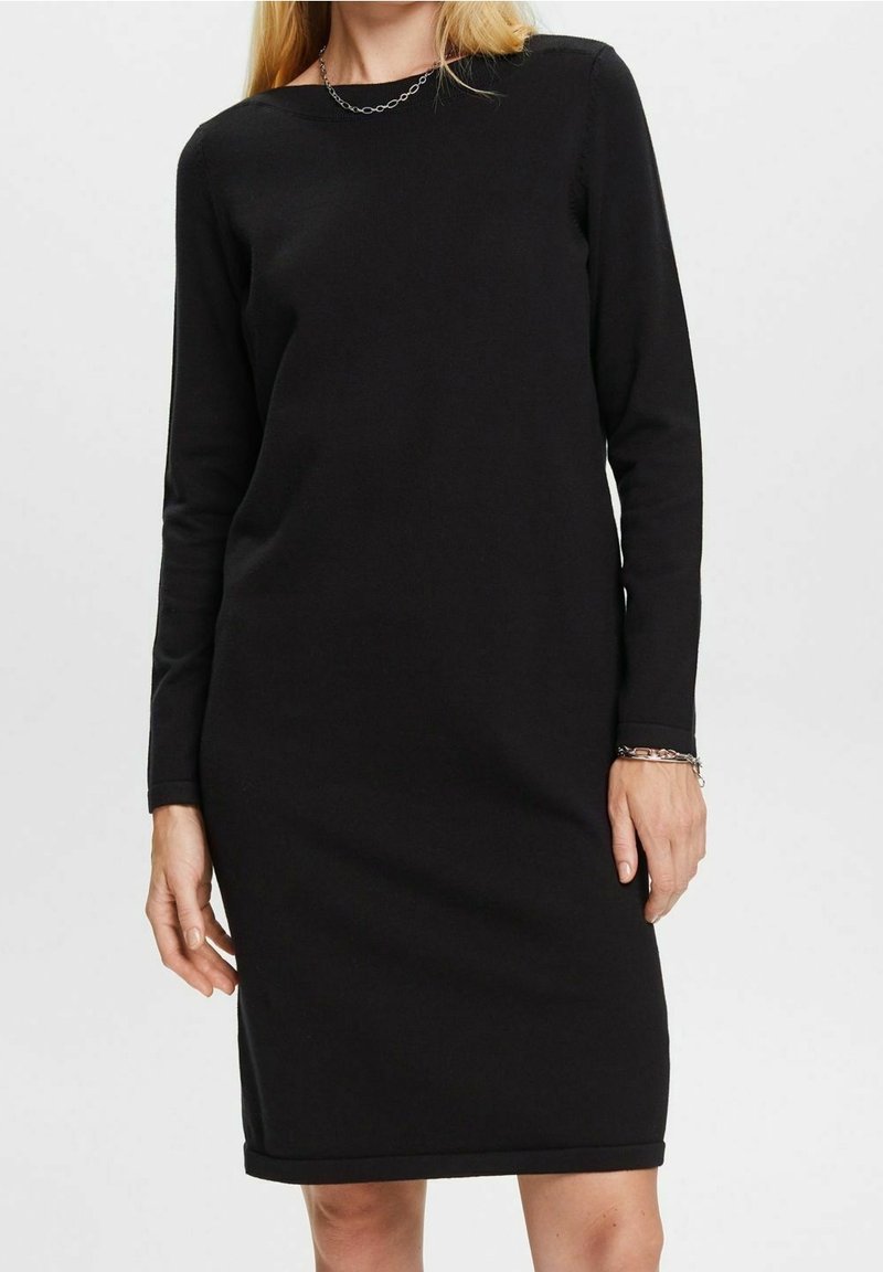 Femme portant une robe noire à manches longues, longueur genou, avec un collier chaîne et un bracelet, debout devant un fond clair uni.