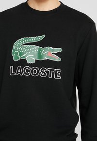 Černá mikina s kapucí s logem zeleného krokodýla a bílými detaily, pod nímž je v tučných bílých písmenách napsáno "LACOSTE".