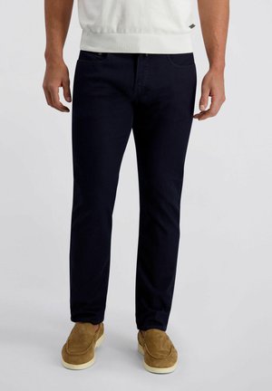 Mann, der dunkel schmal geschnittene Hose, weißen Pullover und beige Wildleder-Slipper trägt und vor einem schlichten grauen Hintergrund steht.