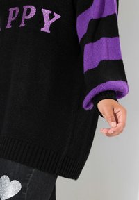Maglione nero a maglia con maniche a strisce viola e nere, caratterizzato da scritte glitterate con la parola "HAPPY". Texture morbida e dettaglio a costine sull'orlo.