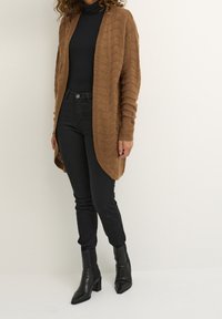 Brauner Strickcardigan mit Kabelmuster, offener Vorderseite, gerippten Ärmeln und weichem Griff, kombiniert mit schwarzen, figurbetonten Jeans und Stiefeletten mit Absatz.