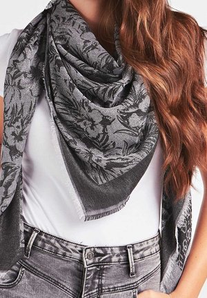 Femme aux longs cheveux ondulés portant un haut blanc, une écharpe grise à motifs enroulée autour du cou et un jean taille haute effet délavé.