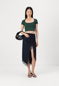 Žalia trumpa rankovė crop top marškinėliai derinami su tamsiai mėlyna nėriniuota maksi sijonu, turinčiu šoninės skeltės ir susiraukšlėjimo dizainą. Modelis laiko juodą rankinę.