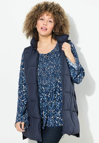 Marinblå pufferväst med hög krage och dragkedja, bärs över en blåmönstrad blus med rund halsringning och långa ärmar.