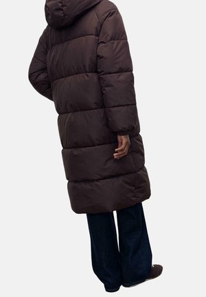 Person mit einem langen, dunkelbraunen Steppmantel mit Kapuze, dunkler, weiter Hose und dunklen Schuhen, stehend vor einem neutralen Hintergrund.