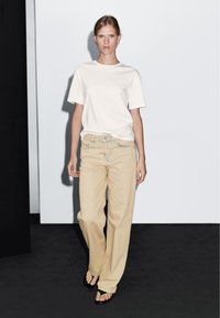 Biała bawełniana koszulka w połączeniu z luźnymi beżowymi dżinsami. Ubranie charakteryzuje się prostym wzornictwem z prostymi spodniami i minimalnymi detalami.