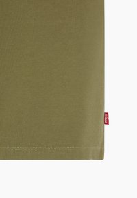 Section de tissu vert olive avec une étiquette rouge de la marque Levi's cousue dans la couture latérale droite près de l'ourlet.