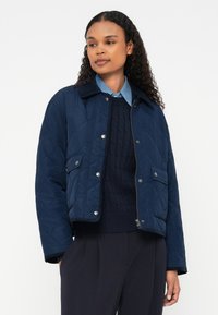 A jaqueta acolchoada azul-marinho apresenta um gola, botões de pressão, dois bolsos frontais e um design curto, usada por cima de um suéter de malha azul-marinho e uma camisa.