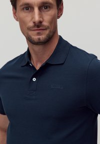 Marinblå polo skjorta i ett texturerat tyg, med en treknapps knäppning, broderad logotyp och kontrasterande ljusblå knappar.