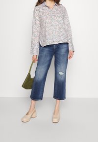 Chemise boutonnée à motif floral avec manches longues, associée à un jean bleu déchiré et des chaussures à talons beiges. Tenant un sac vert.