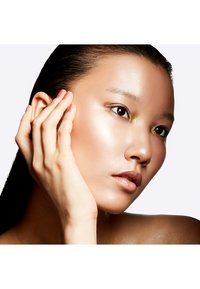 Gros plan sur un visage avec une peau lisse et éclatante, mettant en valeur un léger illuminateur. Cheveux longs et raides et vernis à ongles neutre sur la main touchant la joue.