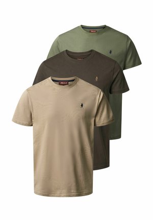 Homme debout, portant un t-shirt beige, un jean foncé et des bottes brunes, souriant avec les bras détendus le long du corps, sur un fond clair uni.