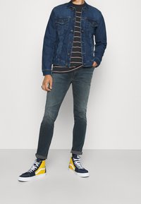 Jeansjacka med två bröstfickor, svartvit randig t-shirt, urtvättade gröna jeans och marinblå-gula högre sneakers.