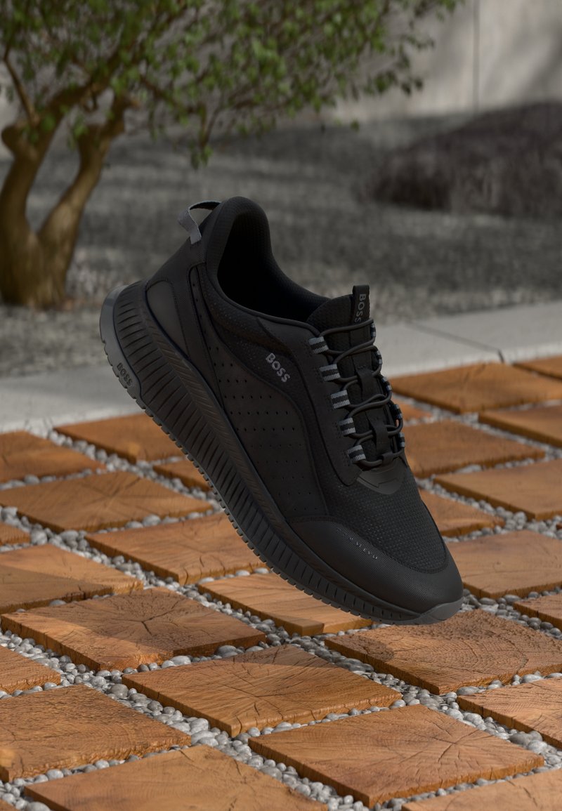 Chaussure de sport noire avec une tige en mesh texturé, des détails perforés et une semelle en caoutchouc. Les lacets sont tissés à travers des œillets renforcés.