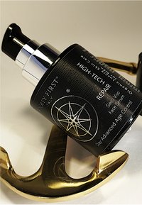 Zwarte cilindervormige fles met een textuurafwerking, chroom pomp en wit logo. Gouden anker-vormig object gedeeltelijk zichtbaar eronder.