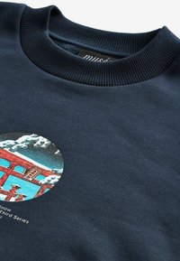 Jūros mėlynumo crewneck tipo džemperis su apvaliu raštu, vaizduojančiu raudoną torii vartus sniege. Lygus paviršius, sukurtas su ripsuotu apykakle ir atspausdintais detalėmis.