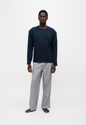 Homme debout pieds nus portant une chemise à manches longues sombre et un pantalon de pyjama rayé de couleur claire sur un fond uni.