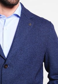 Námořnický modrý blazer z texturované tkaniny s klopovým límcem, vybavený jedním hnědým knoflíkem a mírným leskem, doplněný světle modrou košilí.