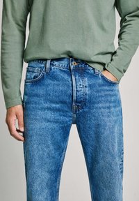 Vaqueros de mezclilla en un tono azul desgastado, con cierre de botón, diseño de cinco bolsillos y corte recto. Combinados con una camisa verde de manga larga.