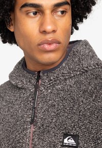 Pull en polaire gris avec col haut, doté d'un zip frontal, d'un tissu texturé et d'une petite étiquette de marque sur la poitrine.