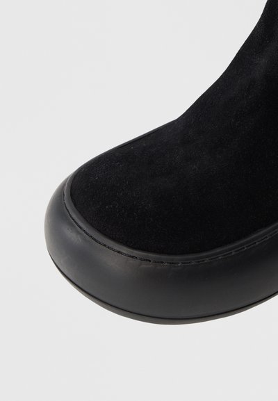 Crna čizma s nubuk gornjištem, zaobljenim vrhom, glatkim crnim gumenim đonom i vidljivim šavovima duž šava.