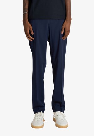 Persona che indossa pantaloni blu navy a gamba dritta, sneakers bianche con suola in gomma e una camicia nera, in piedi davanti a uno sfondo bianco.