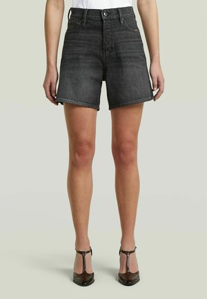 Vrouw draagt zwarte high-waisted denim shorts en zwarte T-strap hakken, staand tegen een effen lichte achtergrond.
