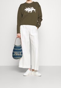 Olivgrön sweatshirt med vit hästtryck, vita linnebyxor med vida ben och en blå vävd väska märkt "POLD." Vita sneakers fulländar looken.