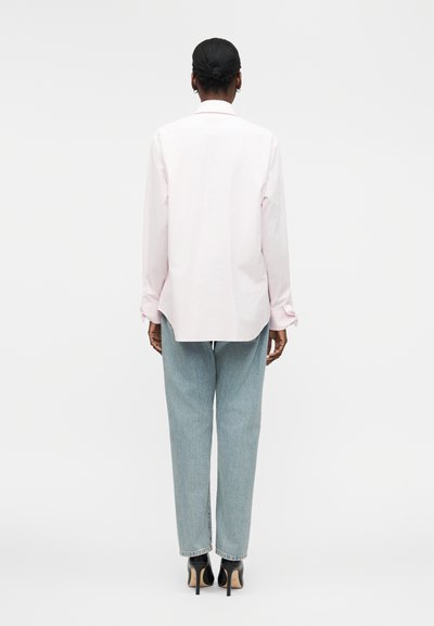 Femme avec les cheveux en chignon, portant une chemise rose pâle à manches longues, un jean bleu clair et des chaussures à talons noirs, debout de dos.