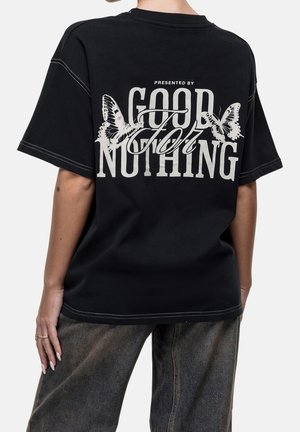 Henkilö, jolla on musta T-paita, jossa valkoinen teksti "Presented by Good for Nothing" ja kaksi perhoskuvitusta selässä.