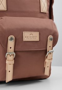 Poche en tissu marron pour sac à dos avec des sangles en cuir beige et des boucles argentées, avec l’étiquette « DOUGHNUT pack your dream ».